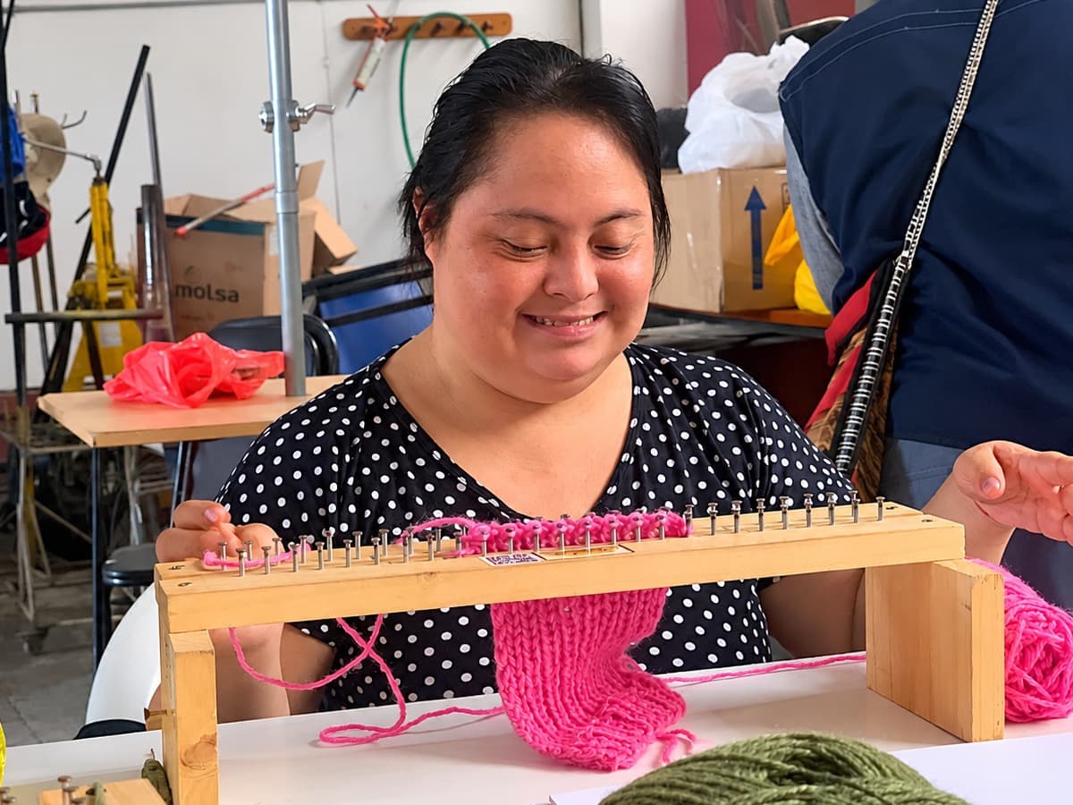 Participante del taller de tejido sonríe mientras teje una pieza rosada en un telar de madera.